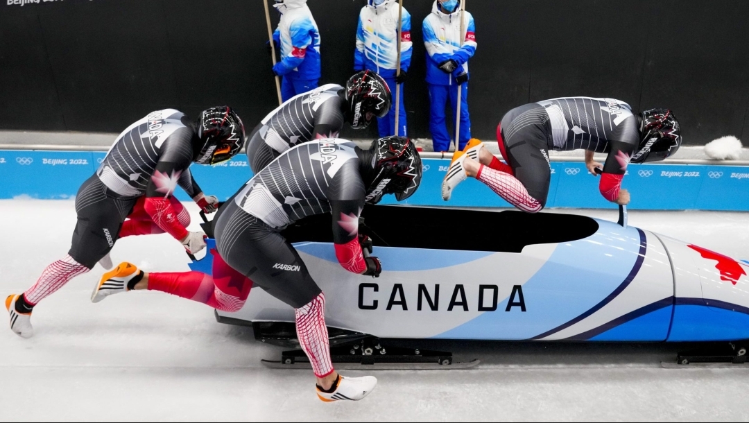 Angle latéral de quatre bobeurs canadiens courant et poussant un bob sur la glace. Le premier en avant est dans les airs, sautant pour entrer dans le bob. Celui-ci est bleu et blanc avec des lignes noires. Sur le côté, on lit Canada et sur le devant du bob, il y a une feuille d'érable rouge.