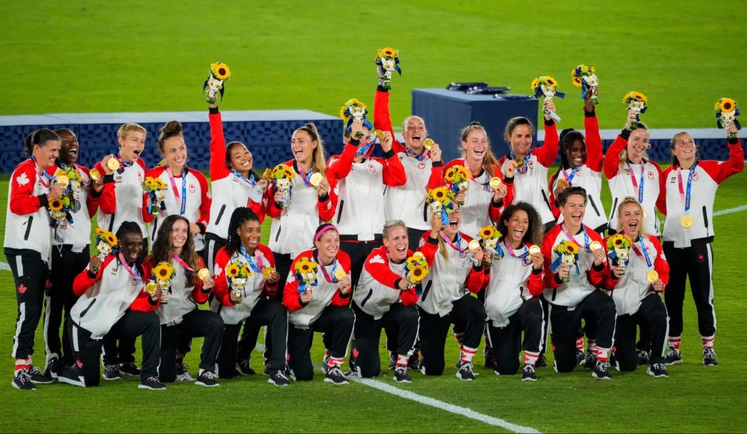 Les membres de l'équipe féminine de soccer sourient, positionnées les unes aux côtés des autres, et lèvent leurs bouquets de fleurs en l'air pour célébrer leur médaille d'or gagnée à Tokyo 2020.