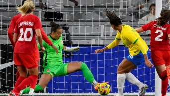 Kailen Sheridan in green uniform blocks the soccer ball with her foot in front of the net