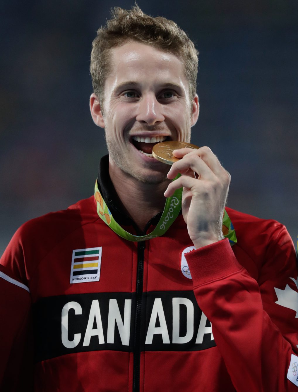 Derek Drouin bites his high jump gold medal at Rio 2016. August 17, 2016. COC Photo/Jason Ransom.