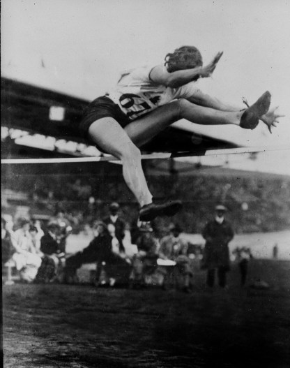 Black and white image of Ethel Catherwood during her high jump. Her left leg is going over the high jump bar with her right about to follow.