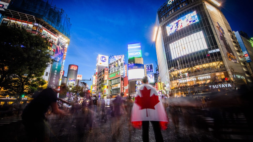 A man holding a Team Canada flag stares out at the crowds walking through Shibuya Crossing.