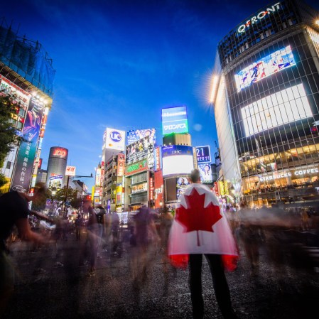 A man holding a Team Canada flag stares out at the crowds walking through Shibuya Crossing.