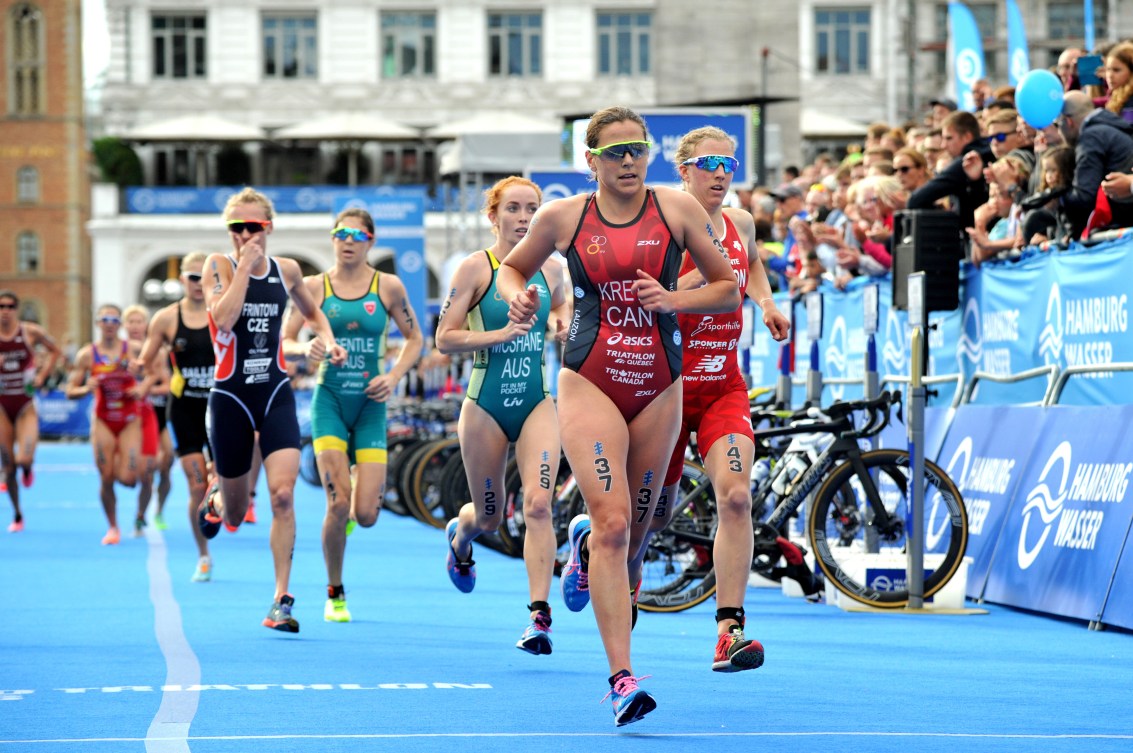 Female triathletes run past spectators (left) and a row of bikes (in front of spectators).