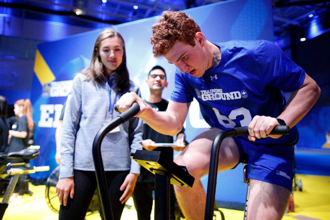 Ian Holmquist rides a stationary bike with two onlookers on the left.