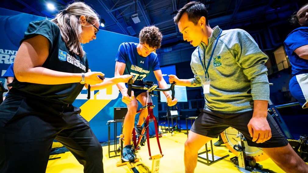 An athlete (centre) rides the sprint bike at the speed station with Patrick Chan (right) cheering him on and a staff (left) keeping track of time.