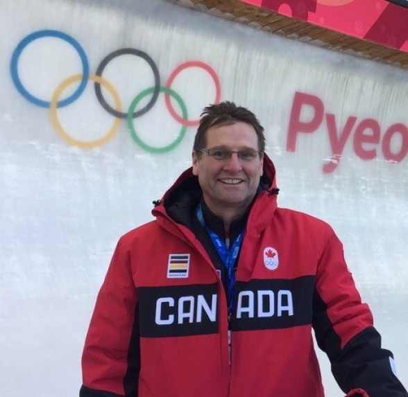 Dr. Mike smiling and standing in front of the bobsleigh track at PyeongChang 2018. He is wearing a red Team Canada winter jacket.