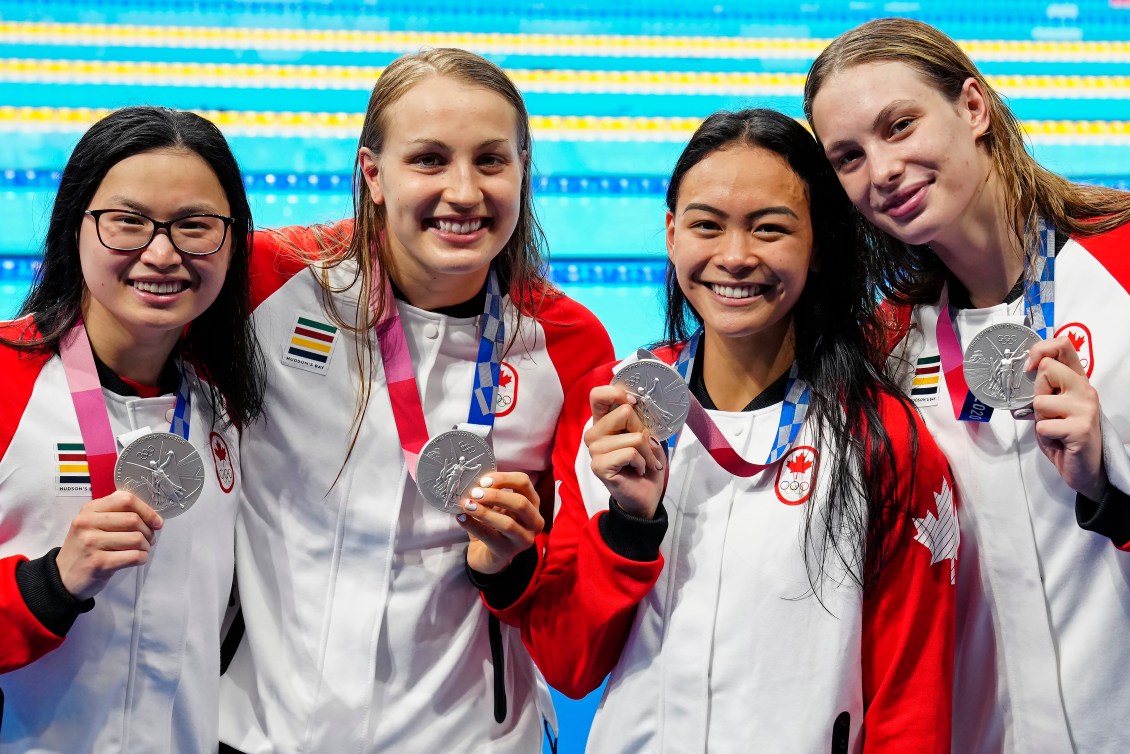 Four Canadian swimmers display their silver medals, standing in front of the Olympic pool.
