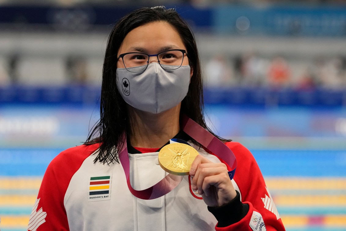 Swimmer Margaret Mac Neil, wearing a Canada jacket and protective mask, holds up her Olympic gold medal.