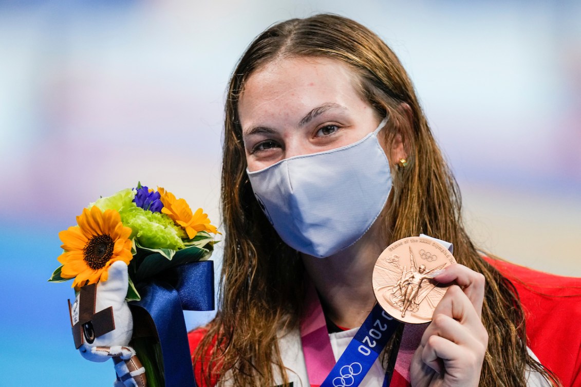 Penny Oleksiak, wearing a Canada jacket and protective mask, holds up a bouquet of flowers and her bronze medal