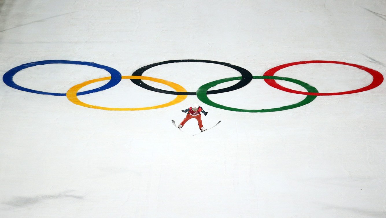 Ski jumper flies through air with Olympic rings in background on the snow