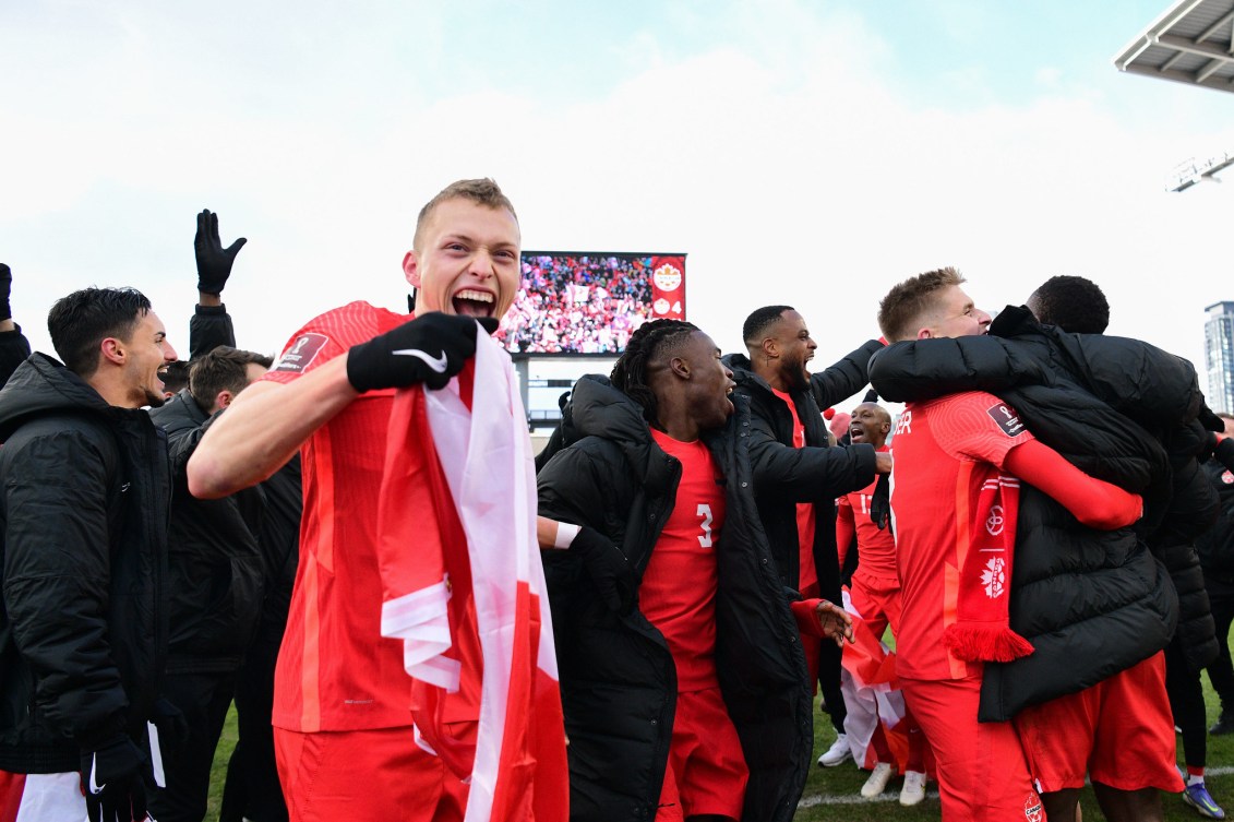 Alistair Johnston holds a Canadian flag and grins while his teammartes celebrate behind him 