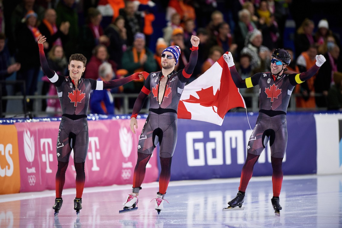 Three speed skaters carry a Canadian flag between them during a victory lap