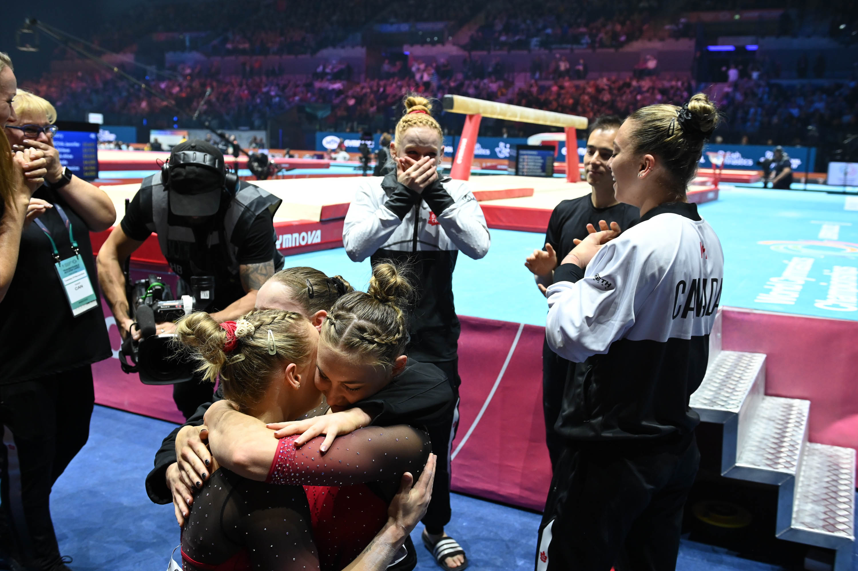 Three gymnasts hug each other on the floor while two others watch in tearful joy