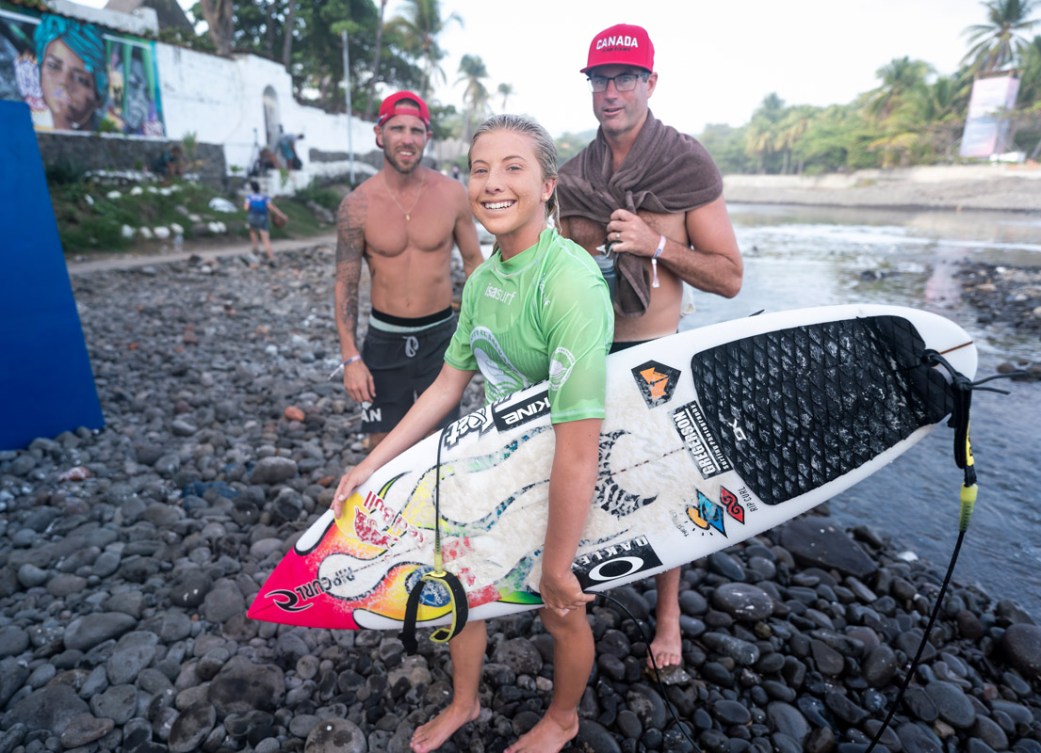 Erin Brooks holds her surfboard while smiling at the camera with teammates