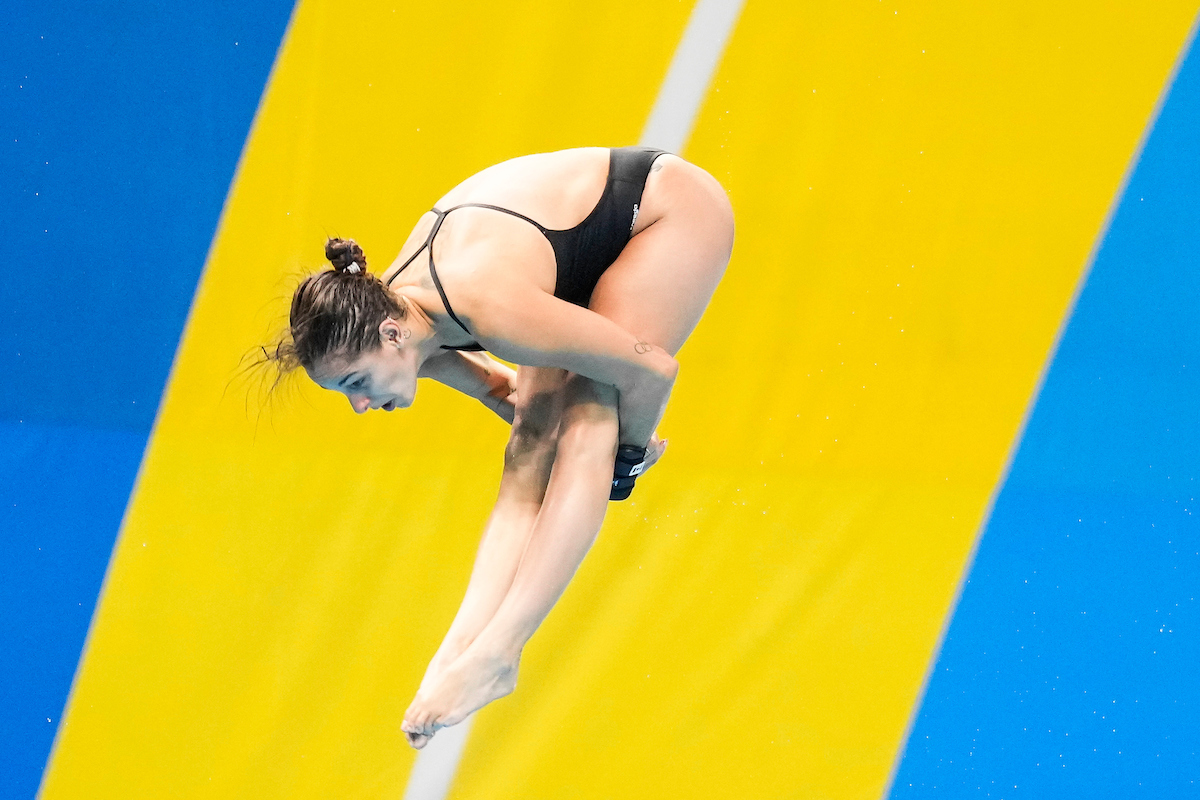 Pamela Ware in a pike position mid dive with a yellow and blue backdrop