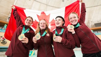 Four women in burgundy jackets wear gold medals and hold a Canadian flag behind them