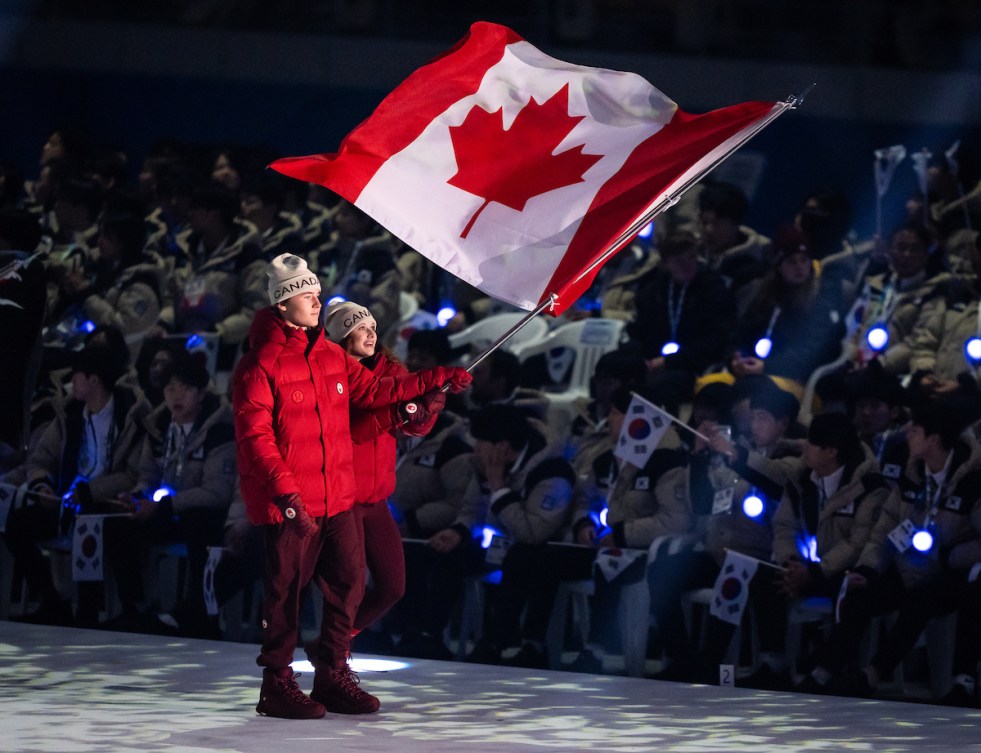 Two Canadian athletes in red jackets carry the Canadian flag 