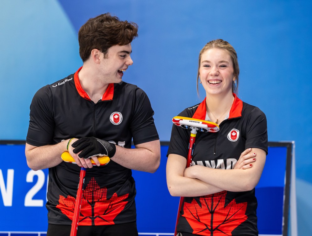 Two Canadian curlers laugh as they stand by the side of the ice sheet 