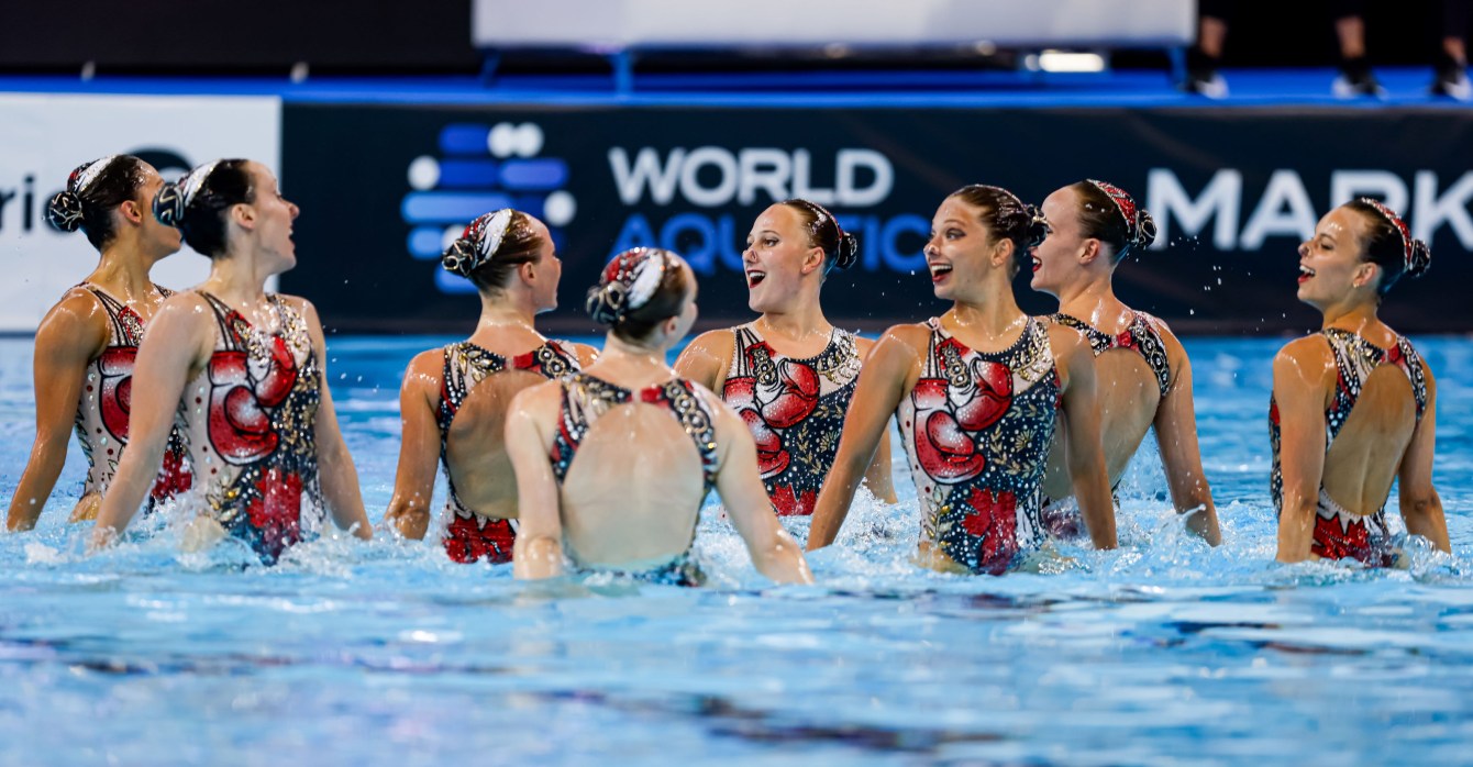 Eight artistic swimmers hold hands as they rise out of the water