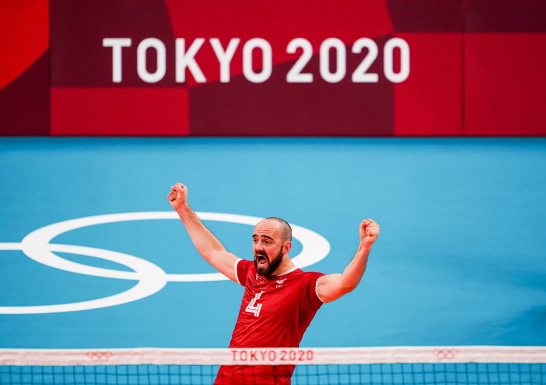 Nicholas Hoag raises both arms in the air in celebration while standing on a blue volleyball court